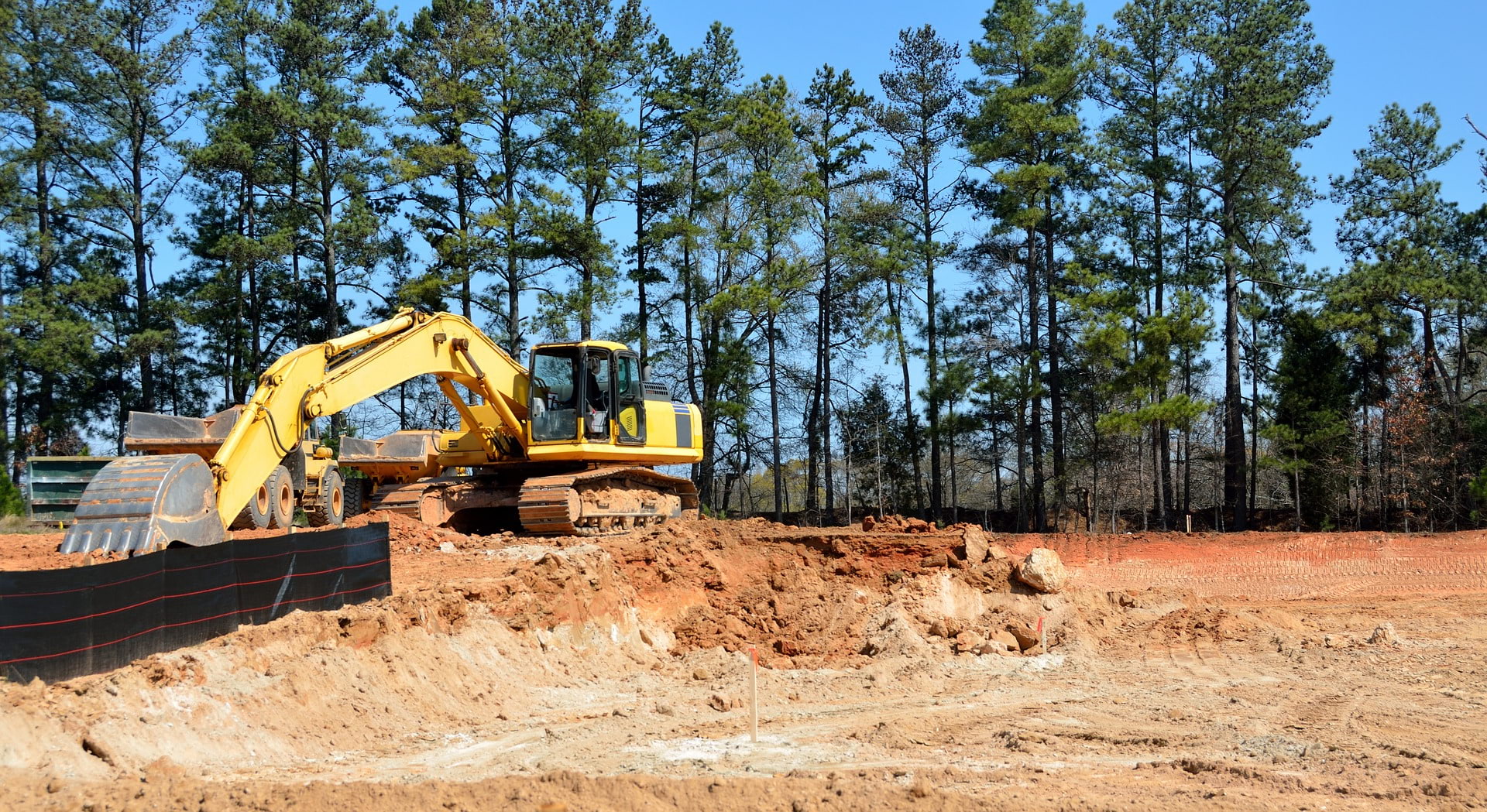 New construction site with excavator digging in red dirt. Backdrop is piney woods and blue sky.