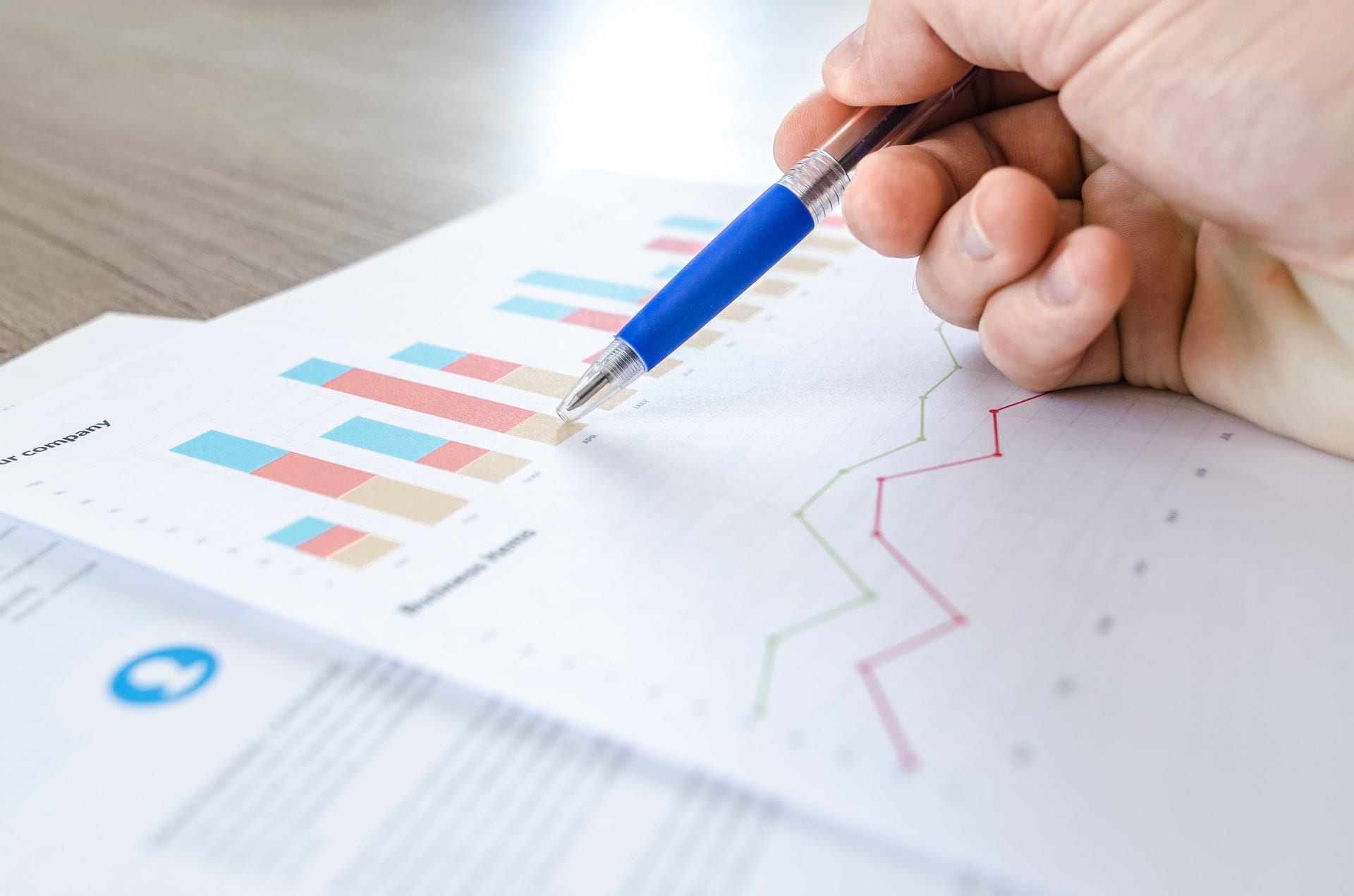 Hand holding a pen pointing to papers with bar charts and scatter-plots on a desk.